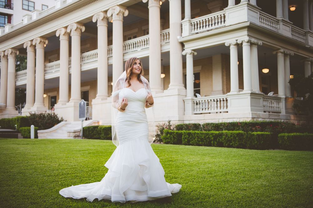 A bride poses in front of a grand white colonial mansion with columns and manicured lawn.