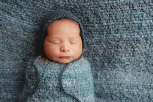 Peaceful newborn baby swaddled in blue knit wrap with matching bonnet on blue blanket.