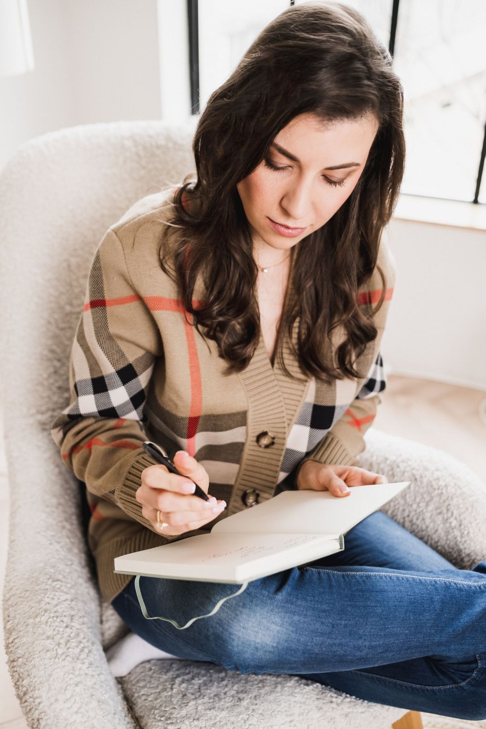Woman in Burberry plaid cardigan writing in notebook while sitting in cozy armchair.