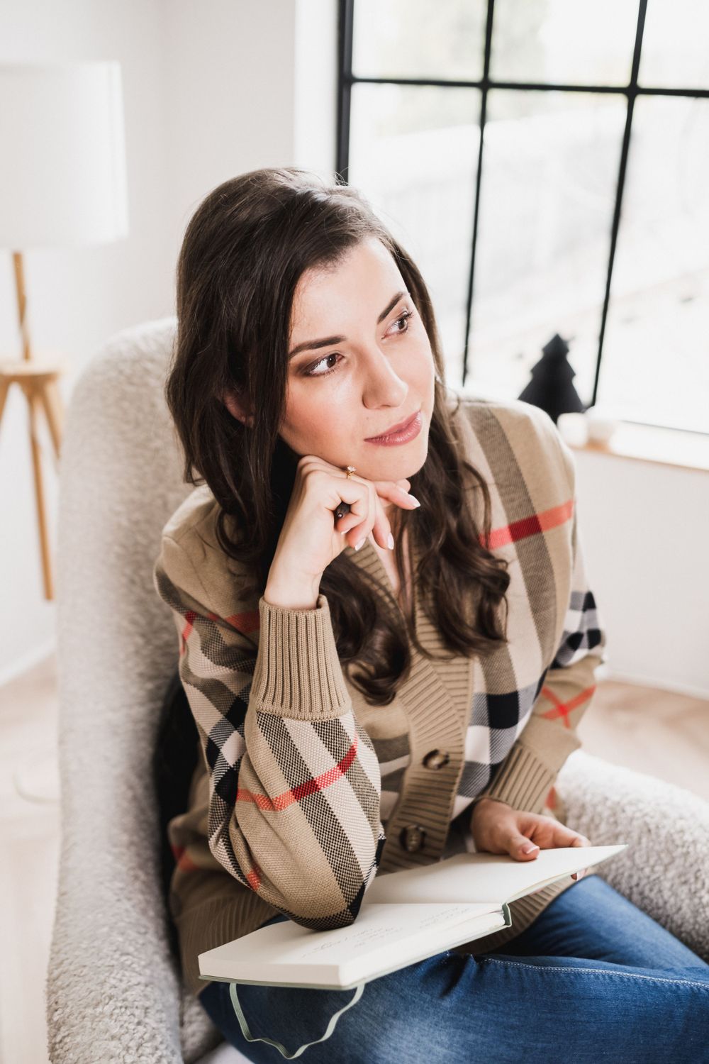 Thoughtful woman in plaid sweater sitting with notebook in bright room.