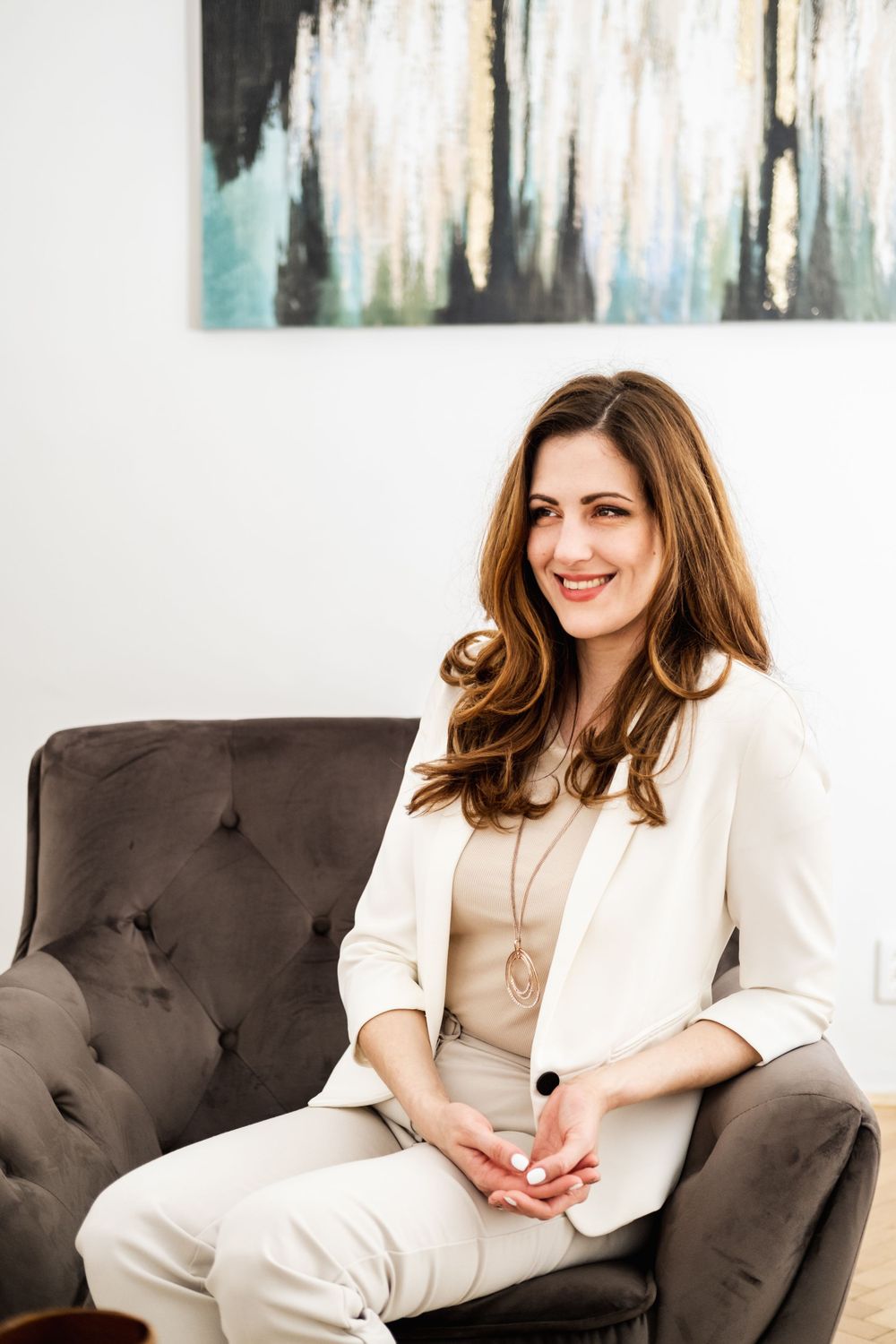 Woman in white blazer sitting in gray armchair with abstract art on wall.