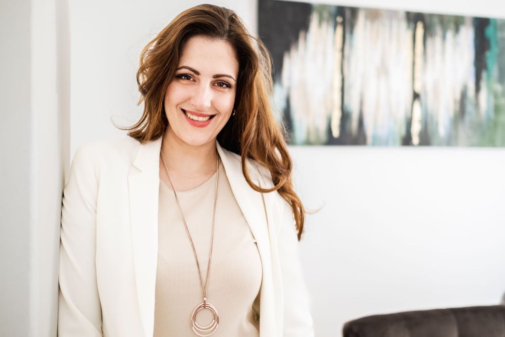 Professional wearing a white blazer and beige top with long pendant necklace in modern office setting.