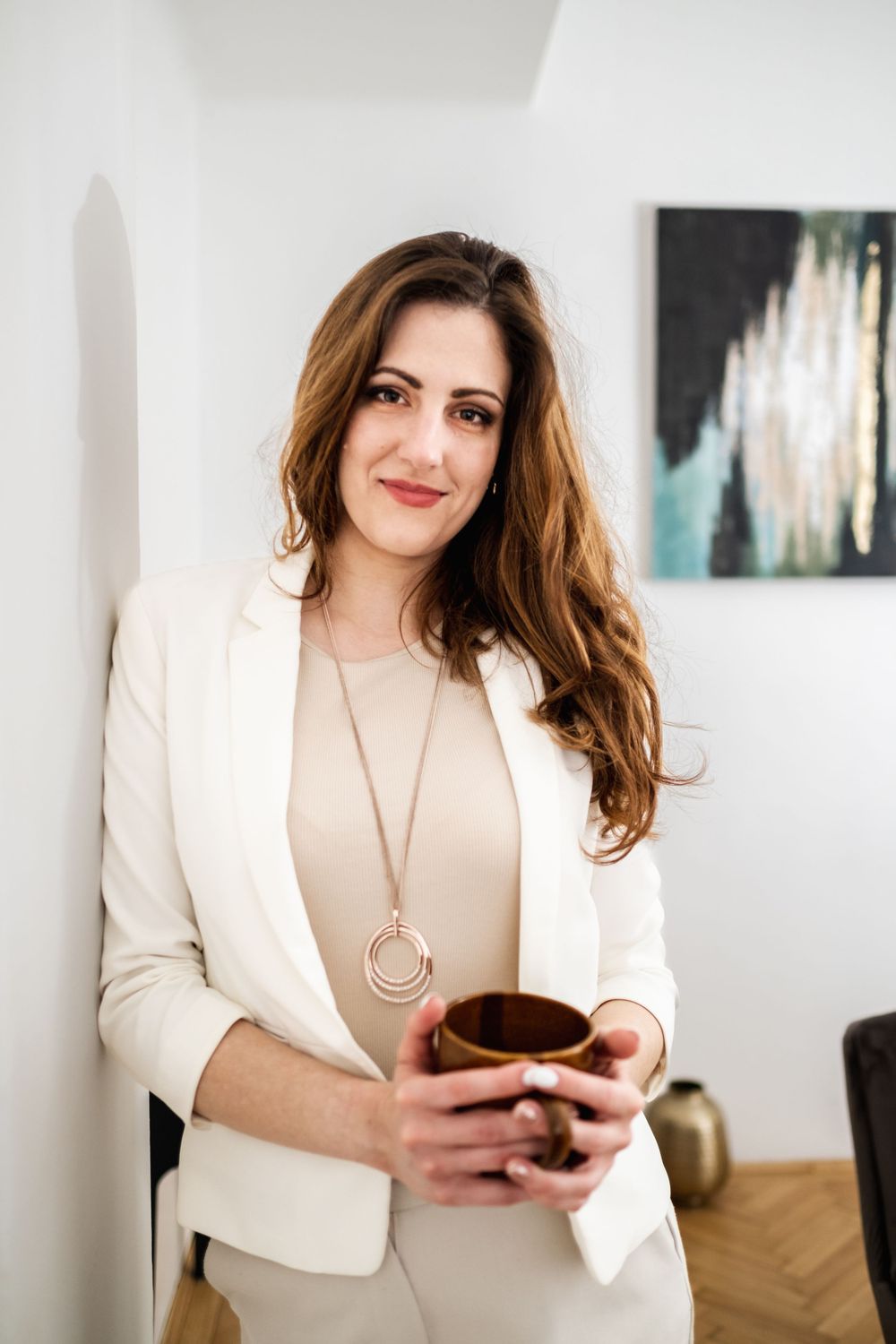 Person in white blazer holding coffee mug against white wall with abstract art.