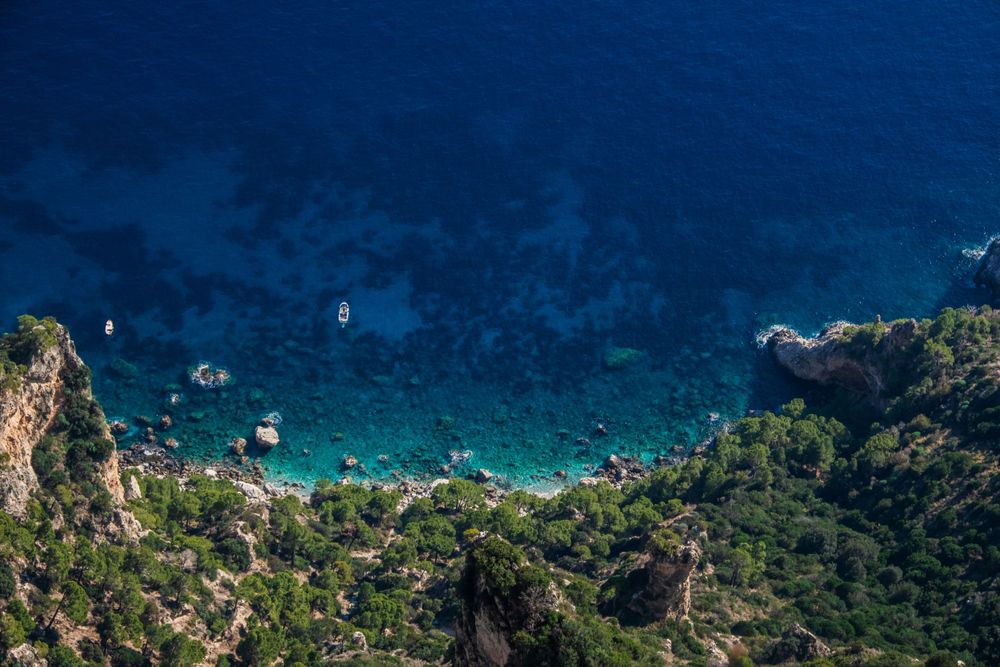 Aerial view of crystal-clear turquoise Mediterranean cove surrounded by rocky cliffs and green trees.