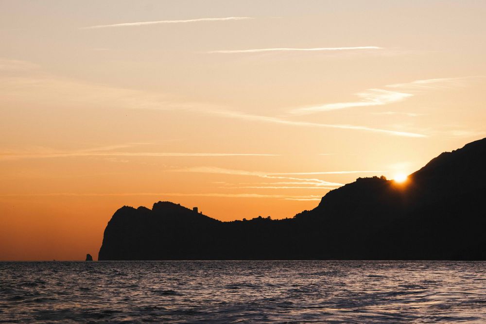 Silhouette of rocky coastal cliffs against a vivid golden orange sunset over calm ocean waters.