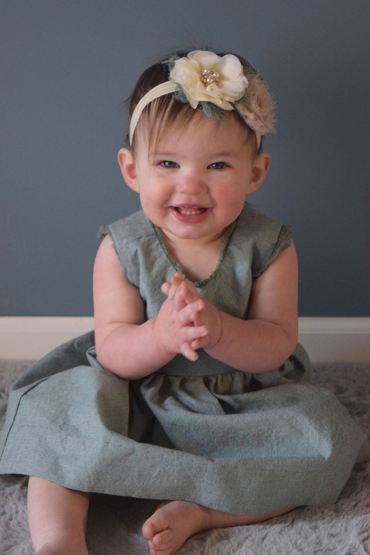 Portrait of a baby in a sage green dress with a floral headband on a gray carpet.