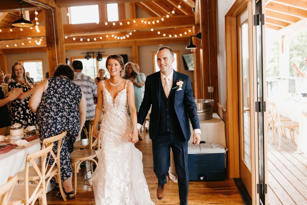 Smiling bride and groom entering rustic barn wedding reception hand in hand through glass doors.