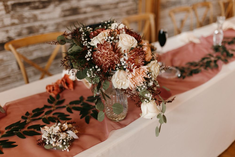 Rustic wedding centerpiece with terracotta dahlias, white roses, and greenery on dusty rose runner.