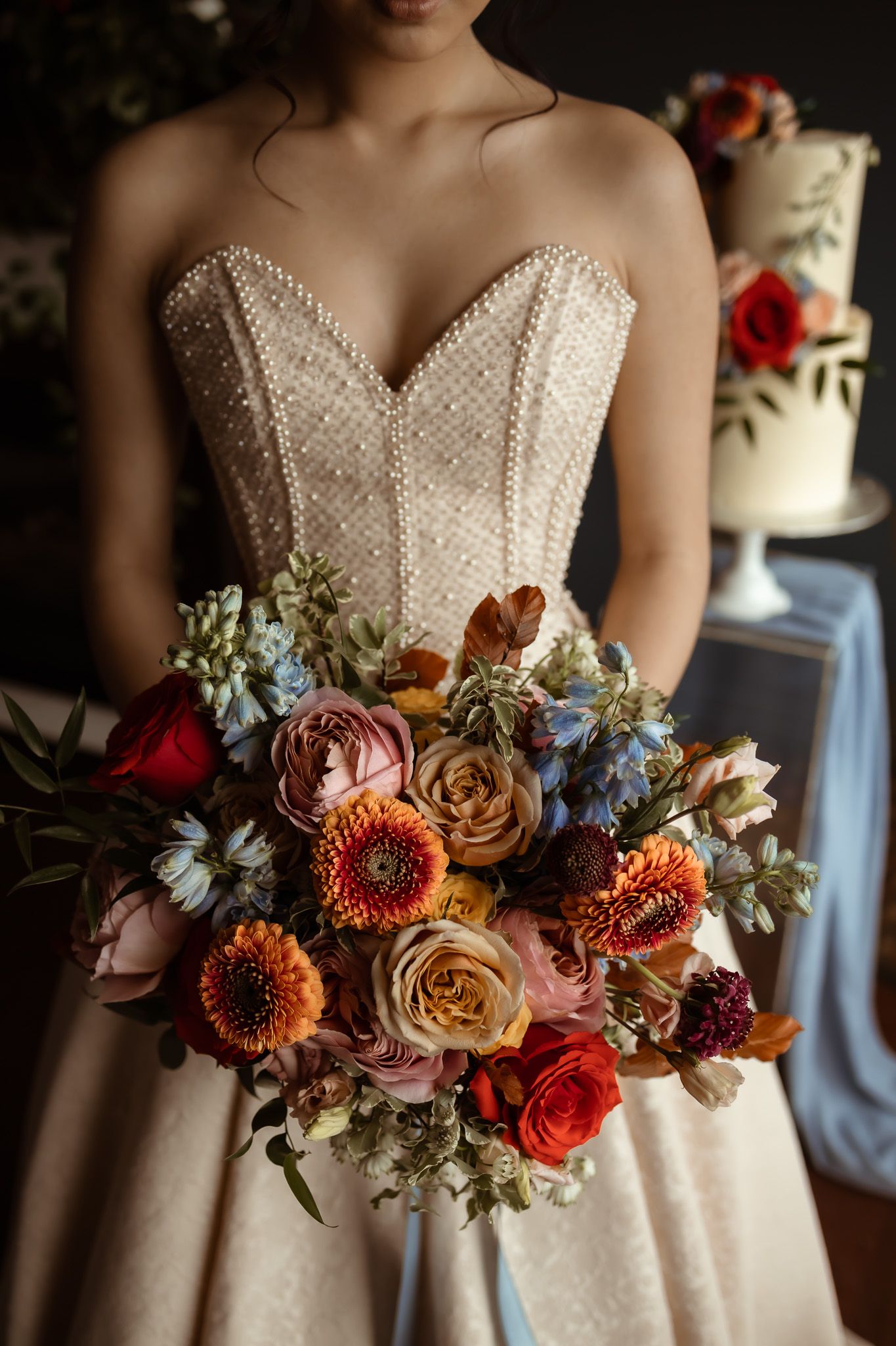 Close-up of a bride in a pearl-beaded wedding dress holding a vibrant autumn bouquet with roses and dahlias.