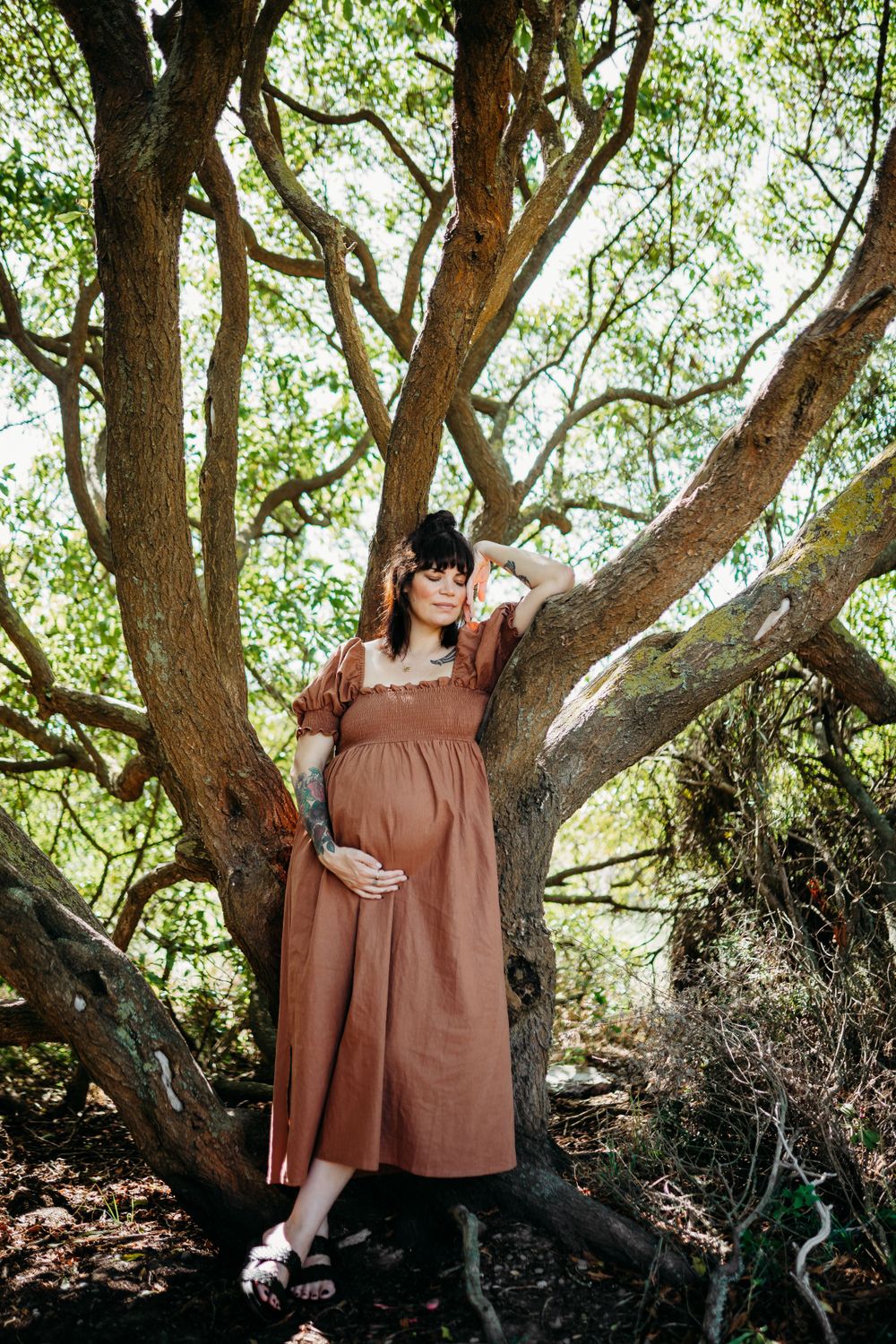Woman in brown prairie dress posing by large tree trunk.