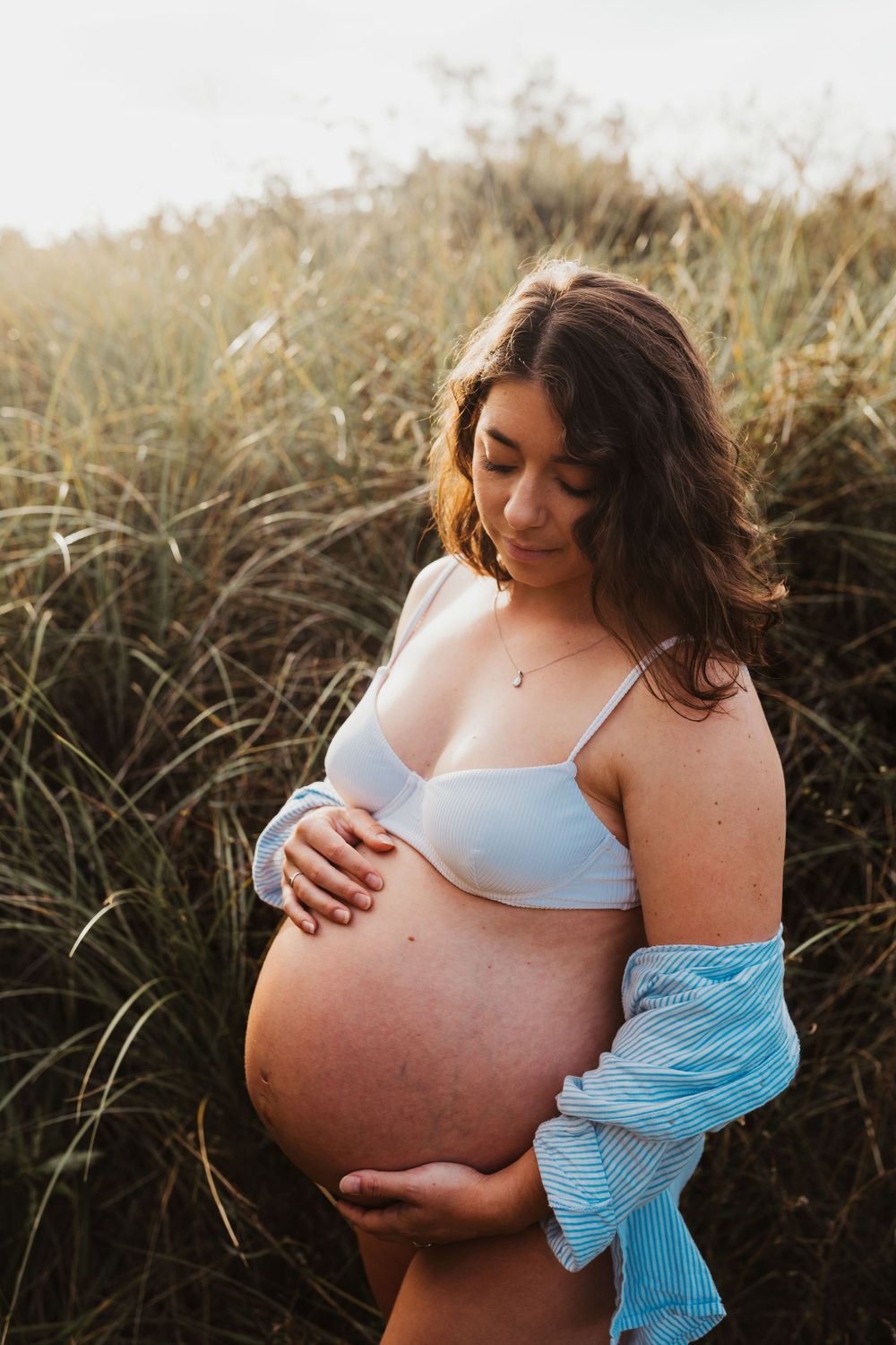 Expectant mother in casual attire standing among tall grass during golden hour.