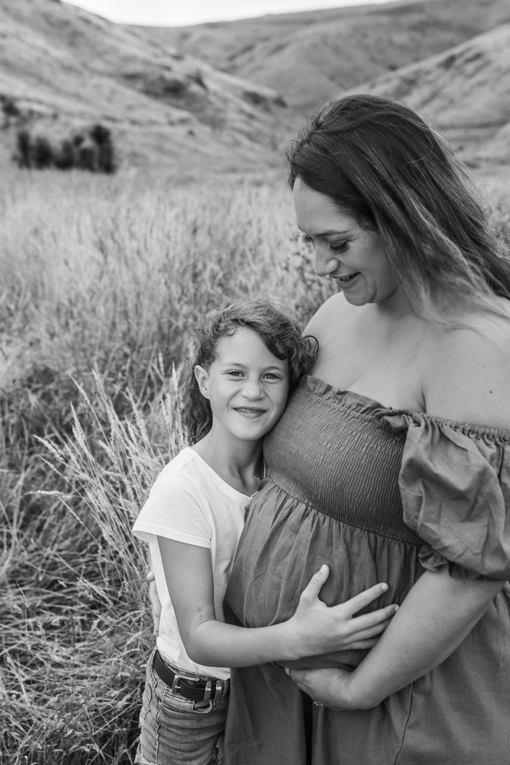 Black and white portrait of expectant mother and child embracing in mountain field.