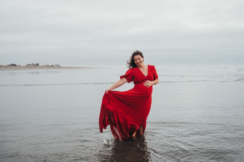 Woman in flowing red dress dancing in shallow ocean waters on a cloudy day.