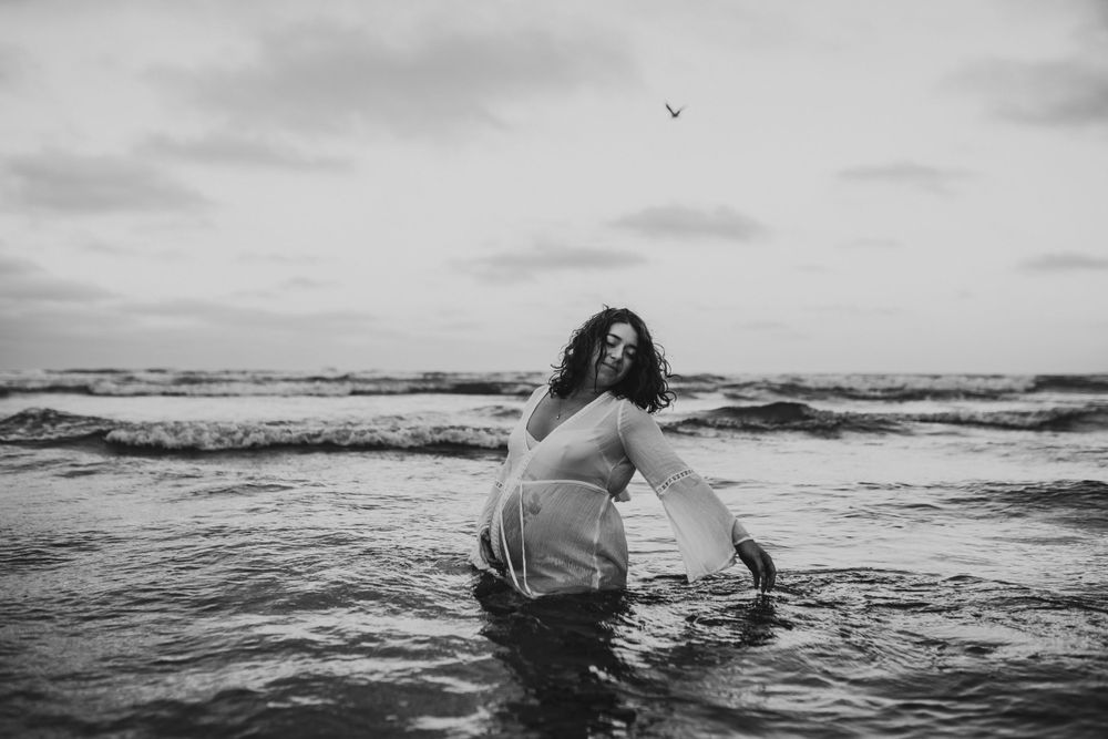 Black and white photo of person in dress wading into ocean waves under cloudy sky.