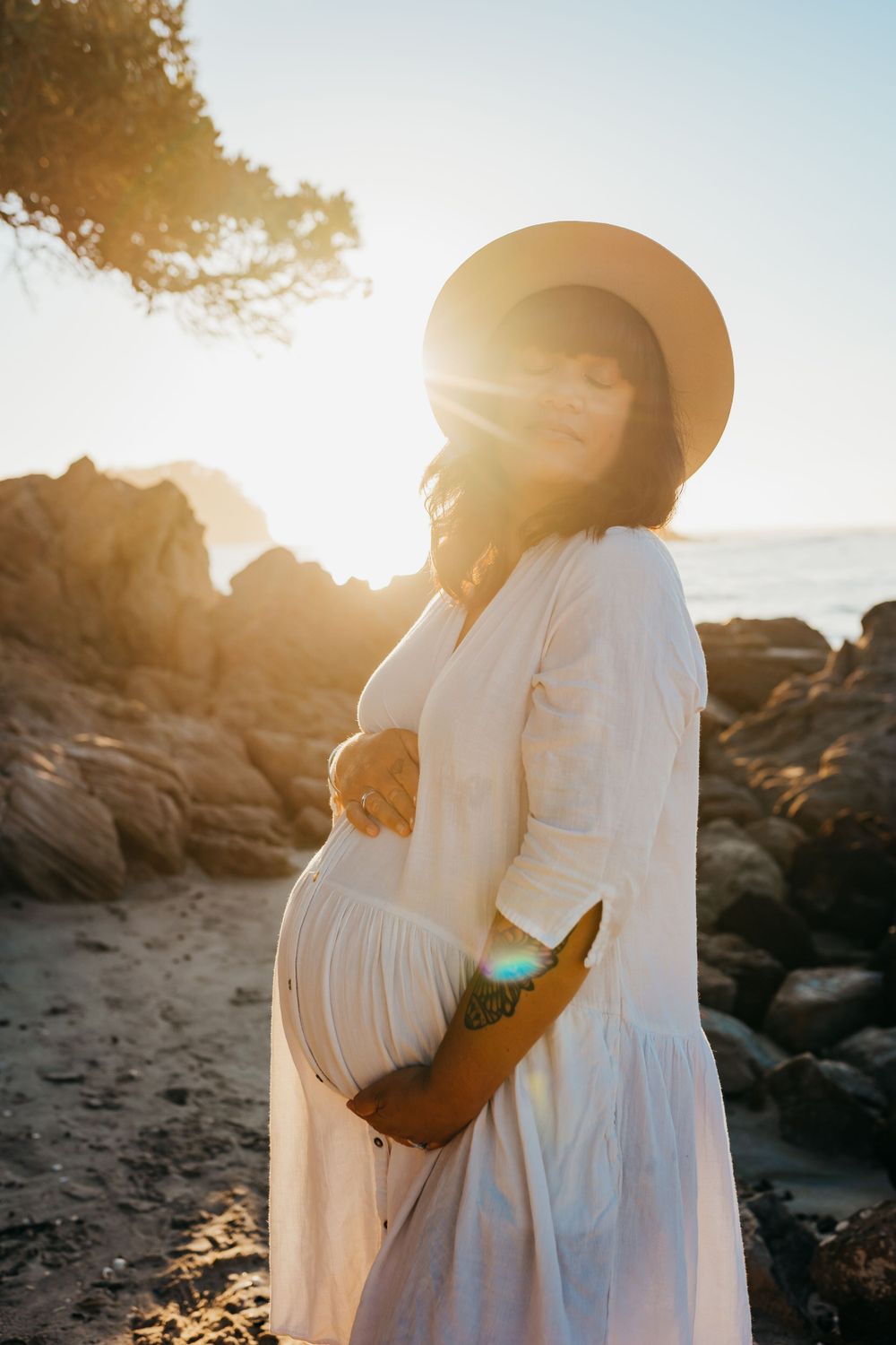 A silhouetted maternity portrait at sunset shows a white dress and sun hat against rocky coastline.