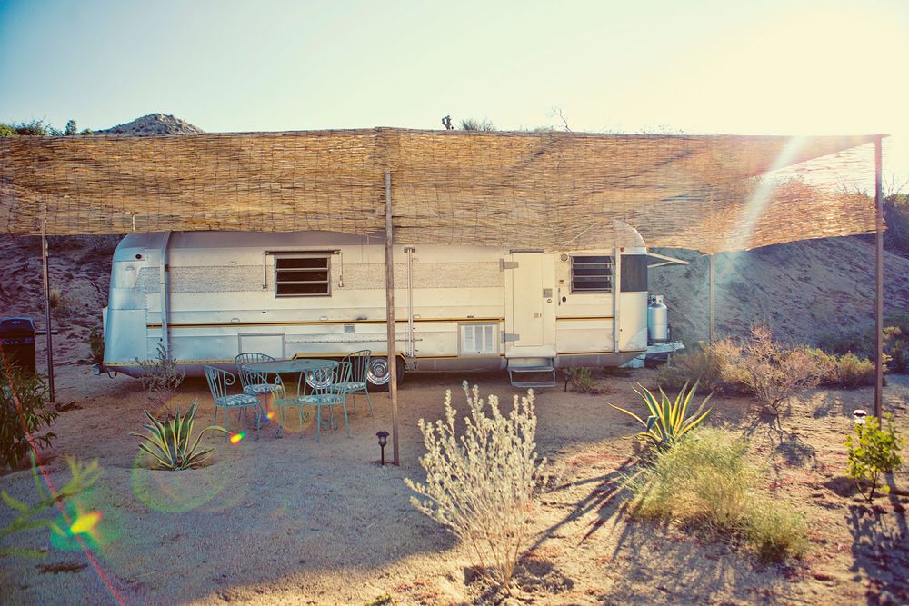 Vintage silver and white travel trailer parked in a sunny desert landscape with sparse desert vegetation.