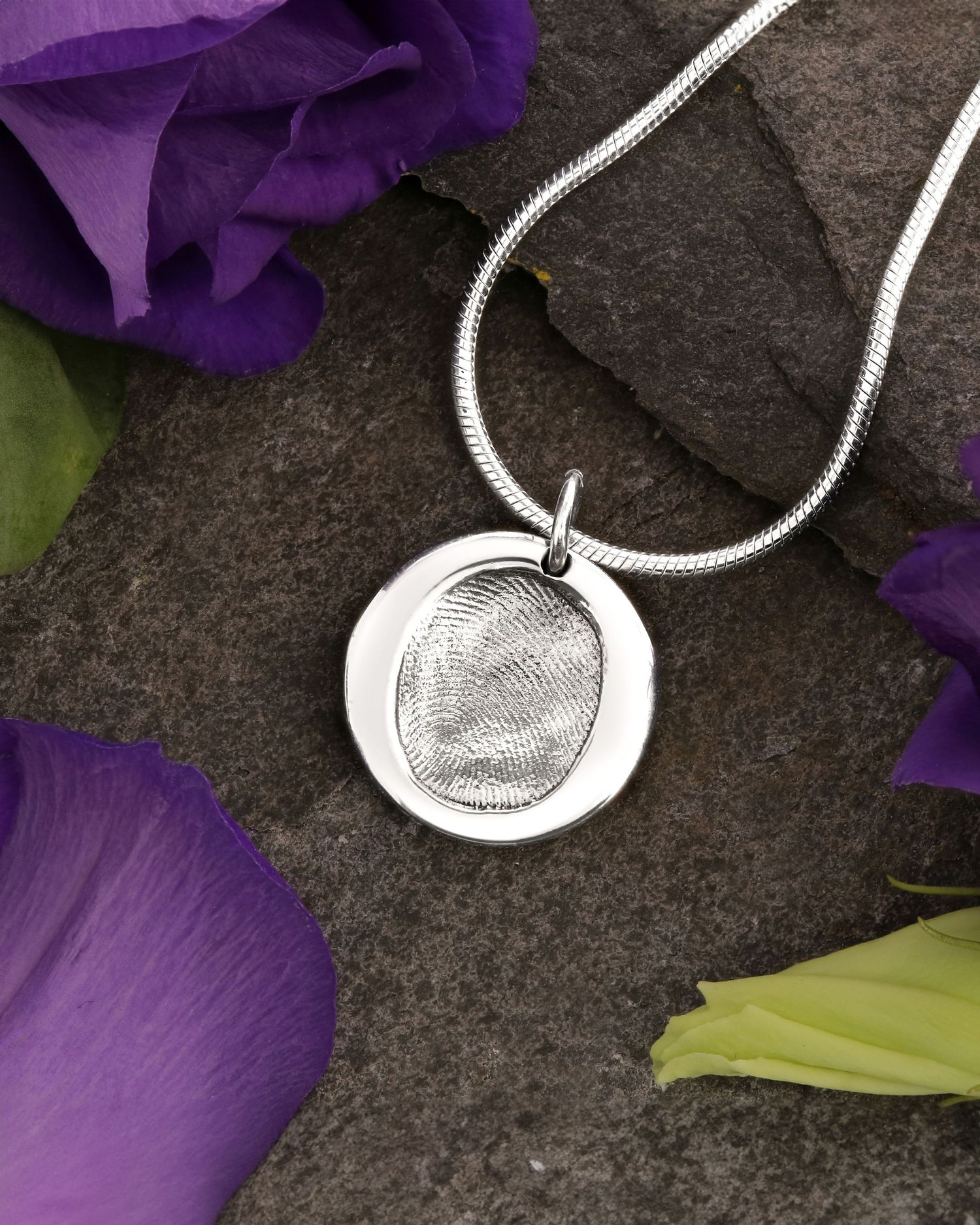Silver fingerprint pendant necklace with snake chain displayed on dark stone with purple flowers.