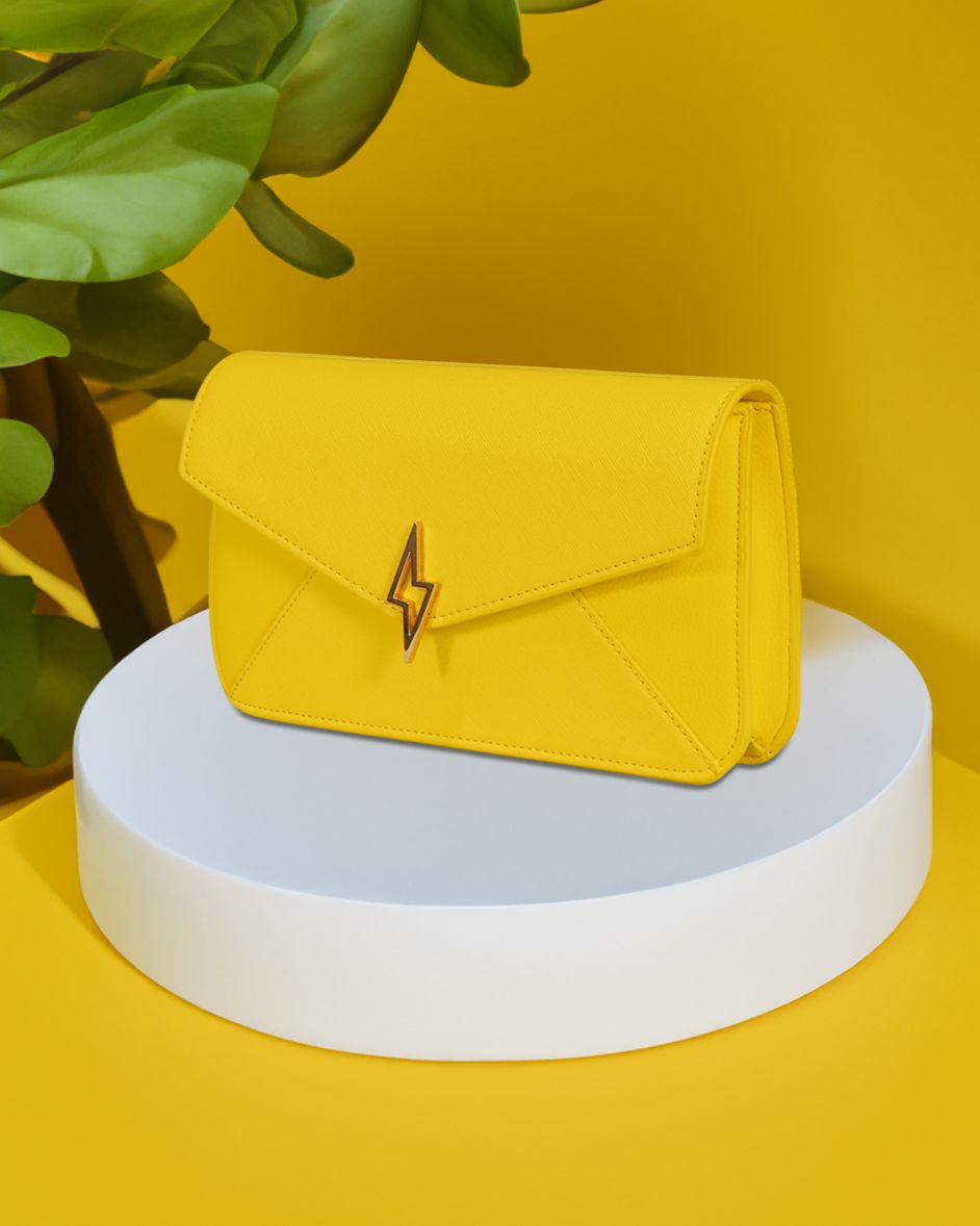 Bright yellow envelope clutch bag with lightning bolt emblem on white pedestal.