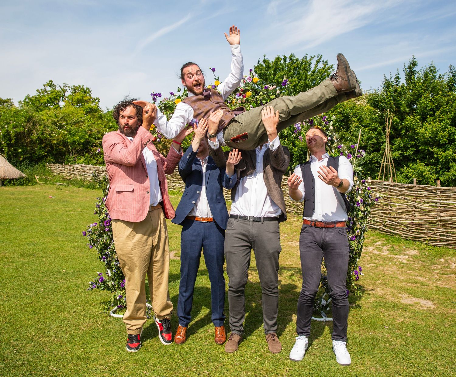 Wedding party members jumping and celebrating on green grass under blue sky.