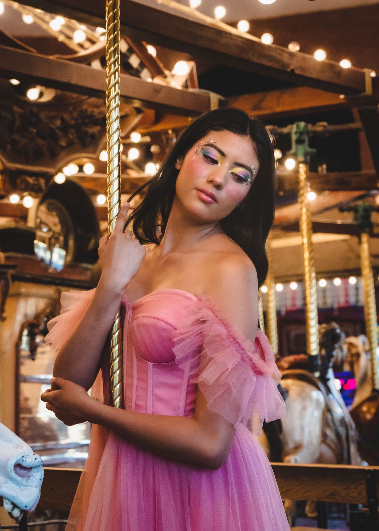 Young woman in a pink tulle off-shoulder gown posing beside a carousel with warm glowing lights.