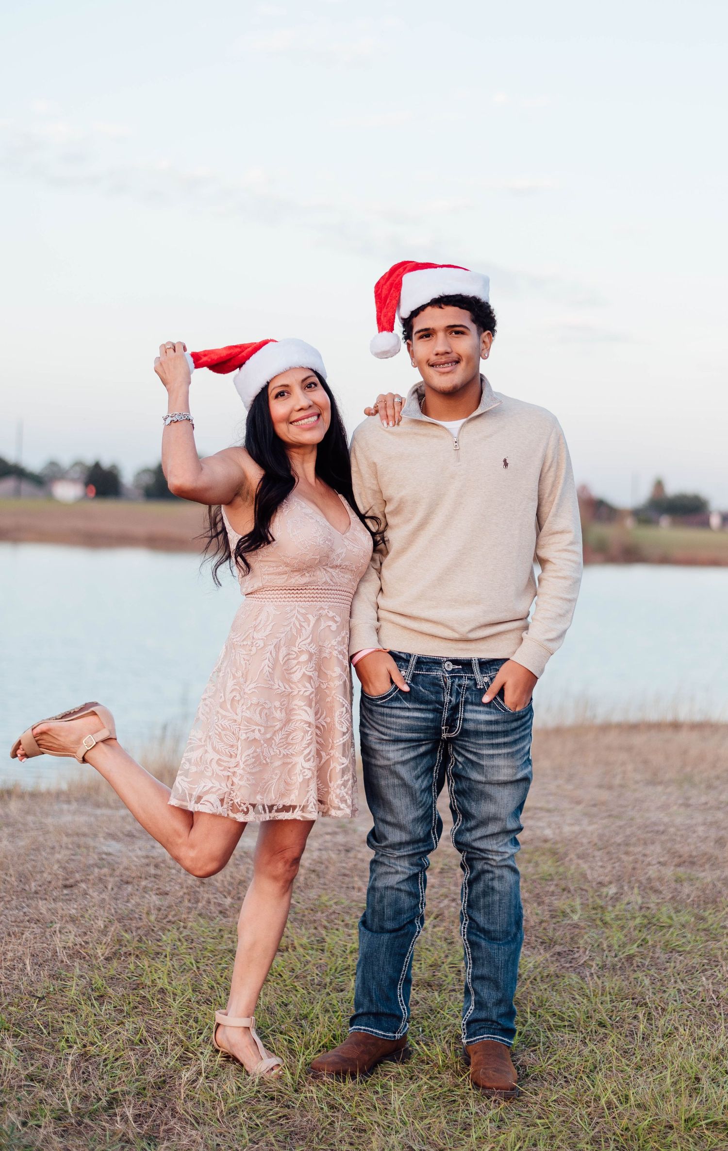 Couple in Santa hats poses playfully by a lake during sunset.