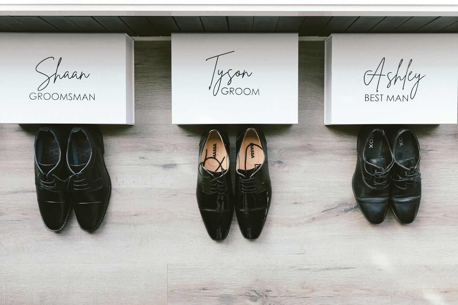 Three pairs of black formal shoes arranged under personalized name cards on light wooden floor.