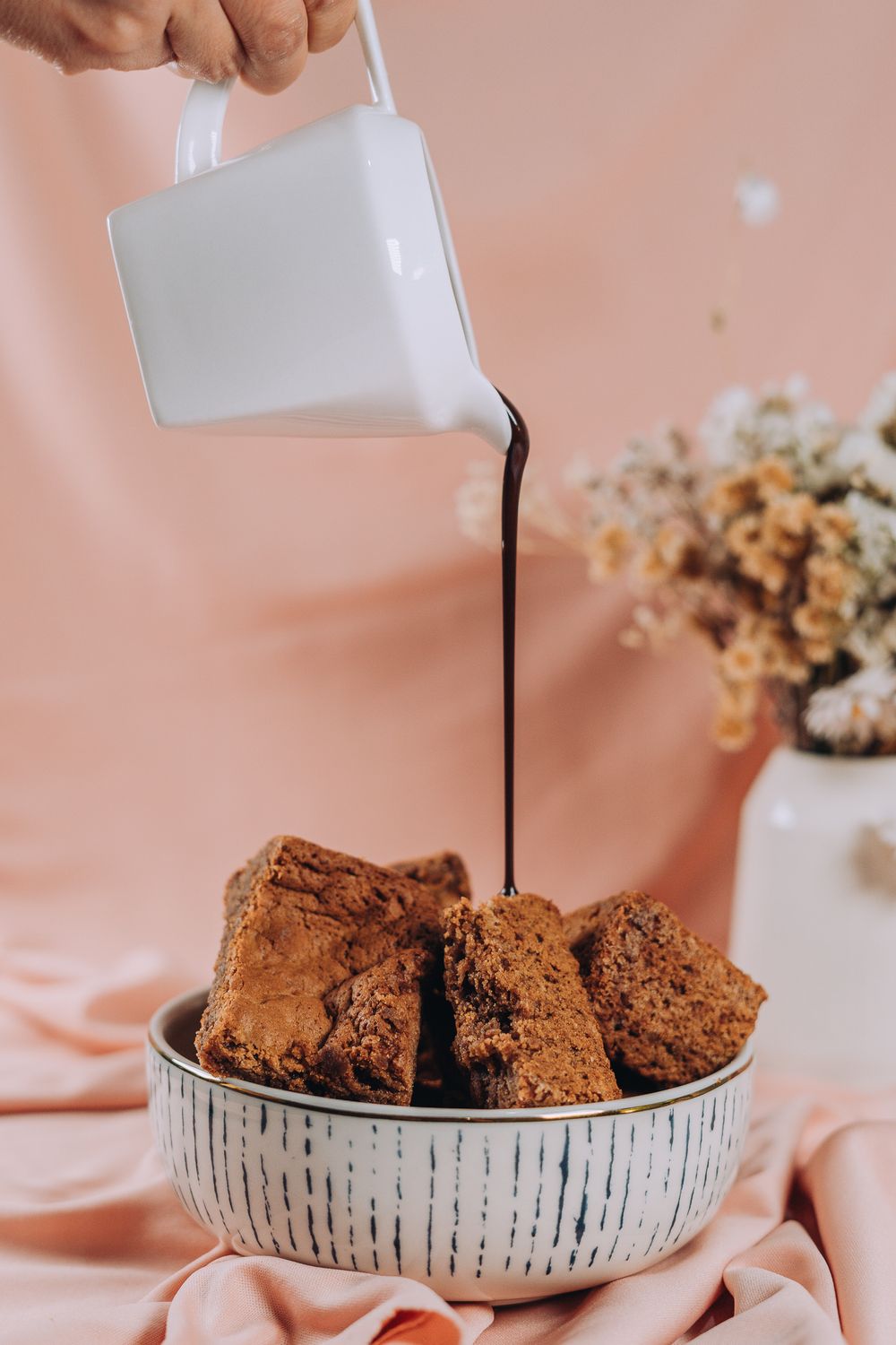 Dark chocolate sauce being poured over brownies in a ceramic bowl against a pink background.