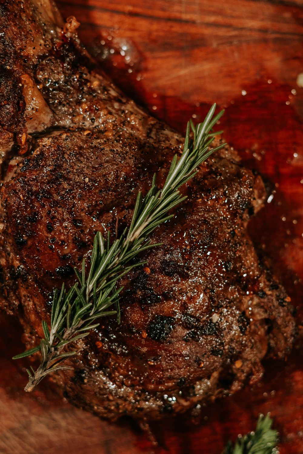 Grilled steak with rosemary sprigs and black pepper crust on wooden surface.