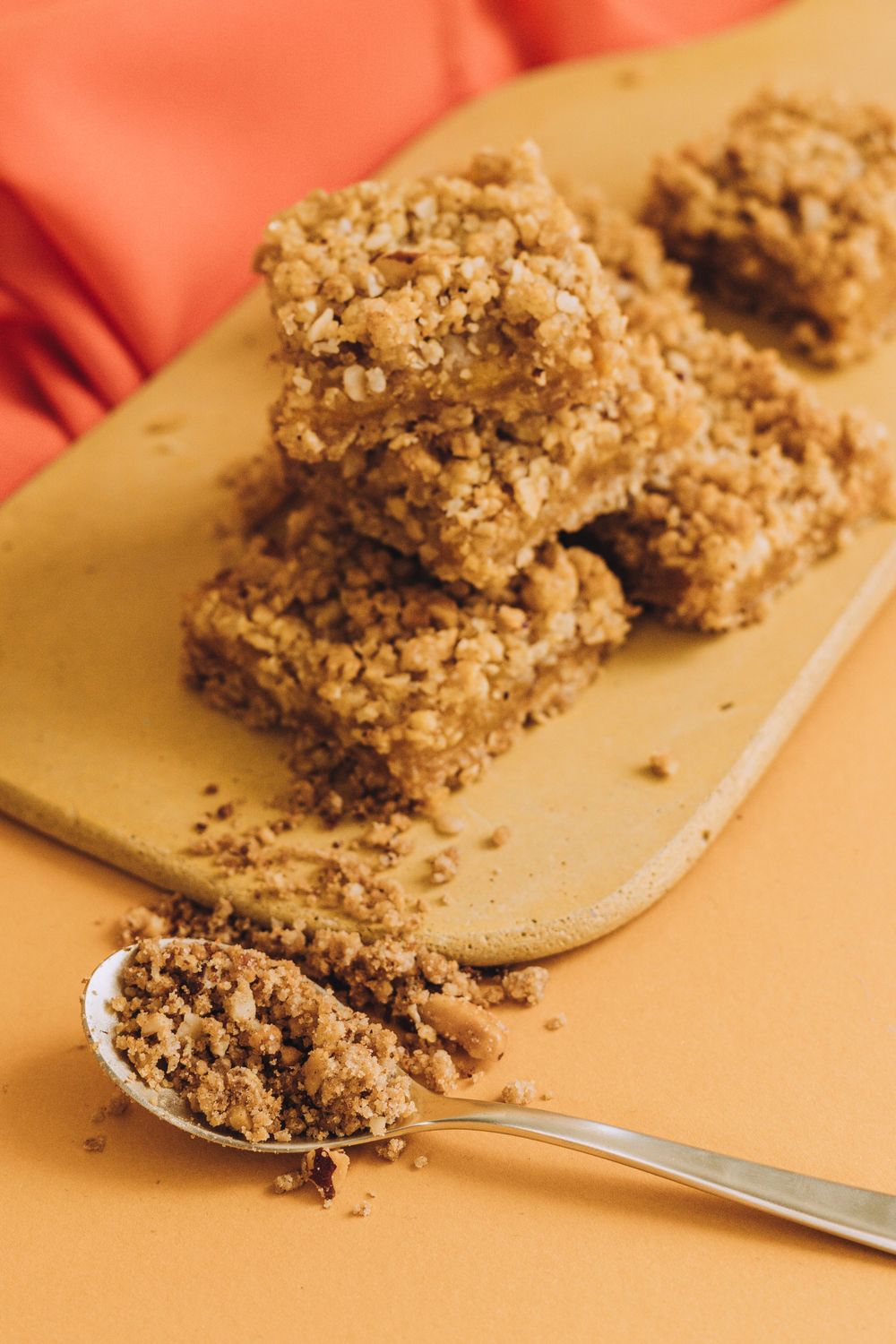 Crumbly oat bars on wooden board with spoonful of mixture on orange background.