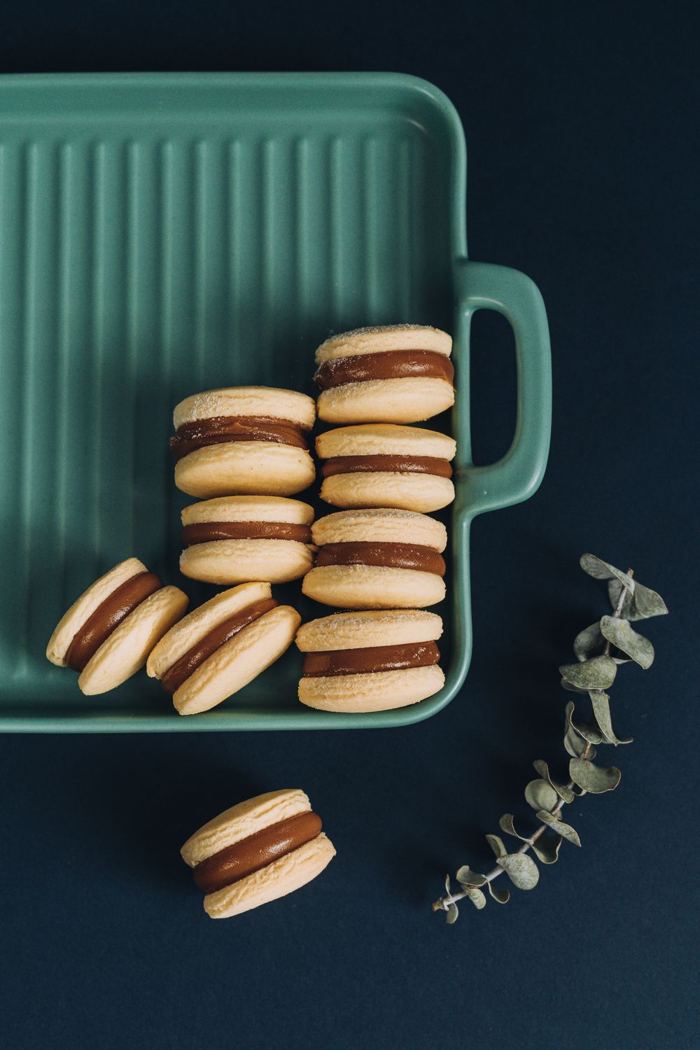 Chocolate sandwich macarons stacked in teal serving dish with eucalyptus sprig.