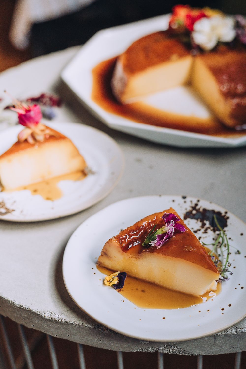 Three slices of caramel flan garnished with edible flowers on white plates.