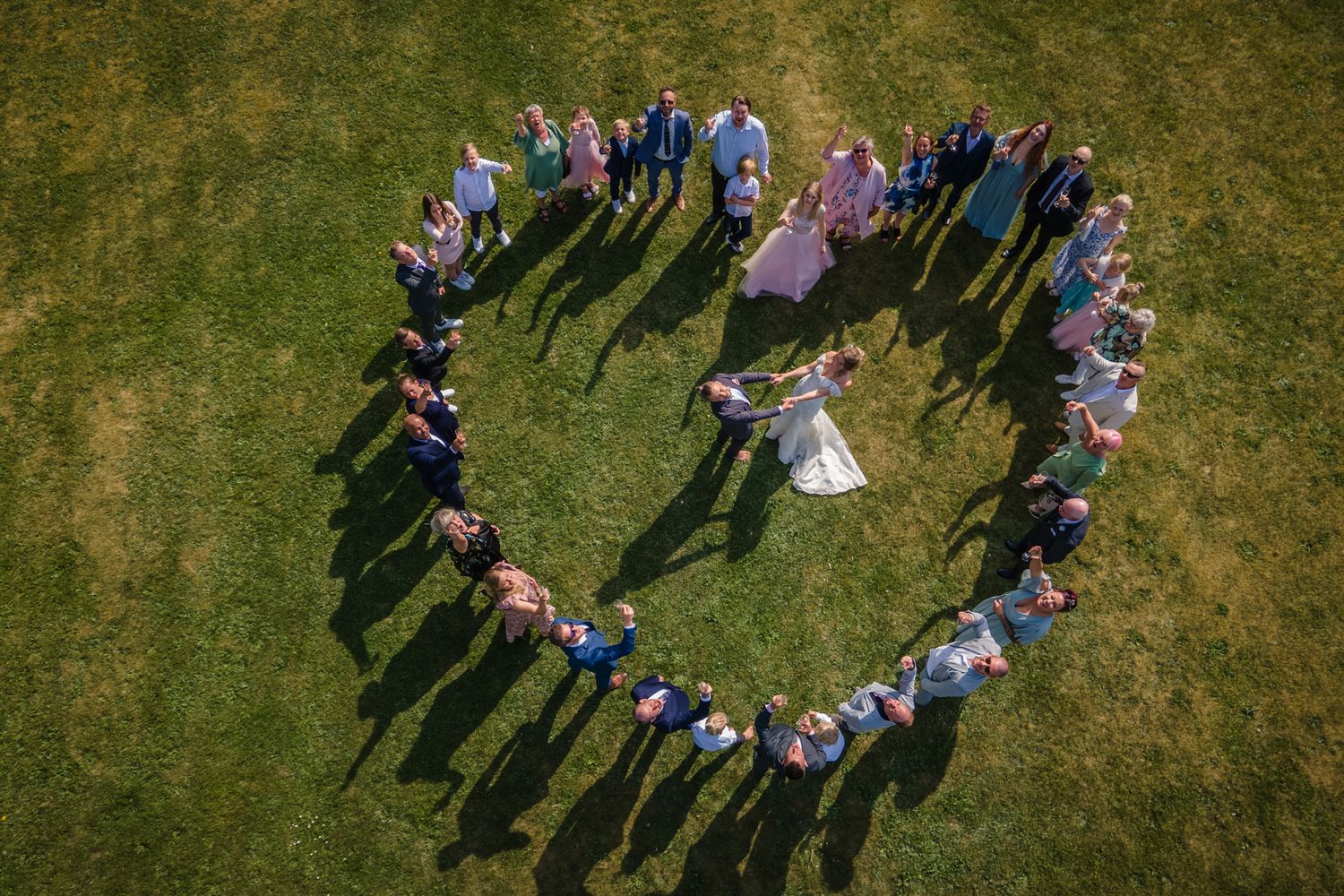 Bröllopsfotografering på Hjelmsjöborg Örkelljunga. Gruppbild av bröllopsgäster som står runt brudpar i ett hjärta.