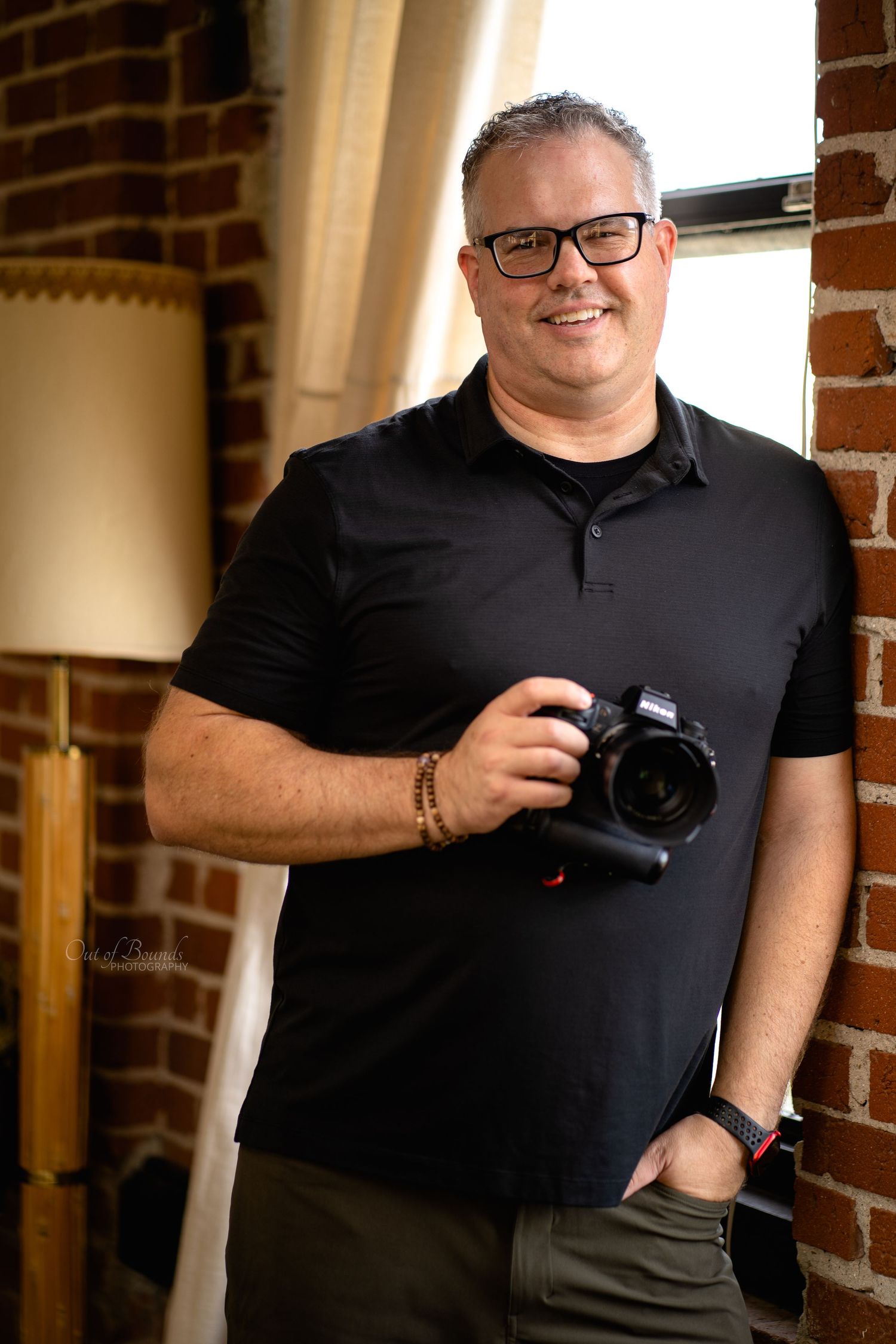Professional photographer holding a mirrorless Nikon Z9 camera posing against exposed brick wall in casual black polo. Tucker Maxfield
