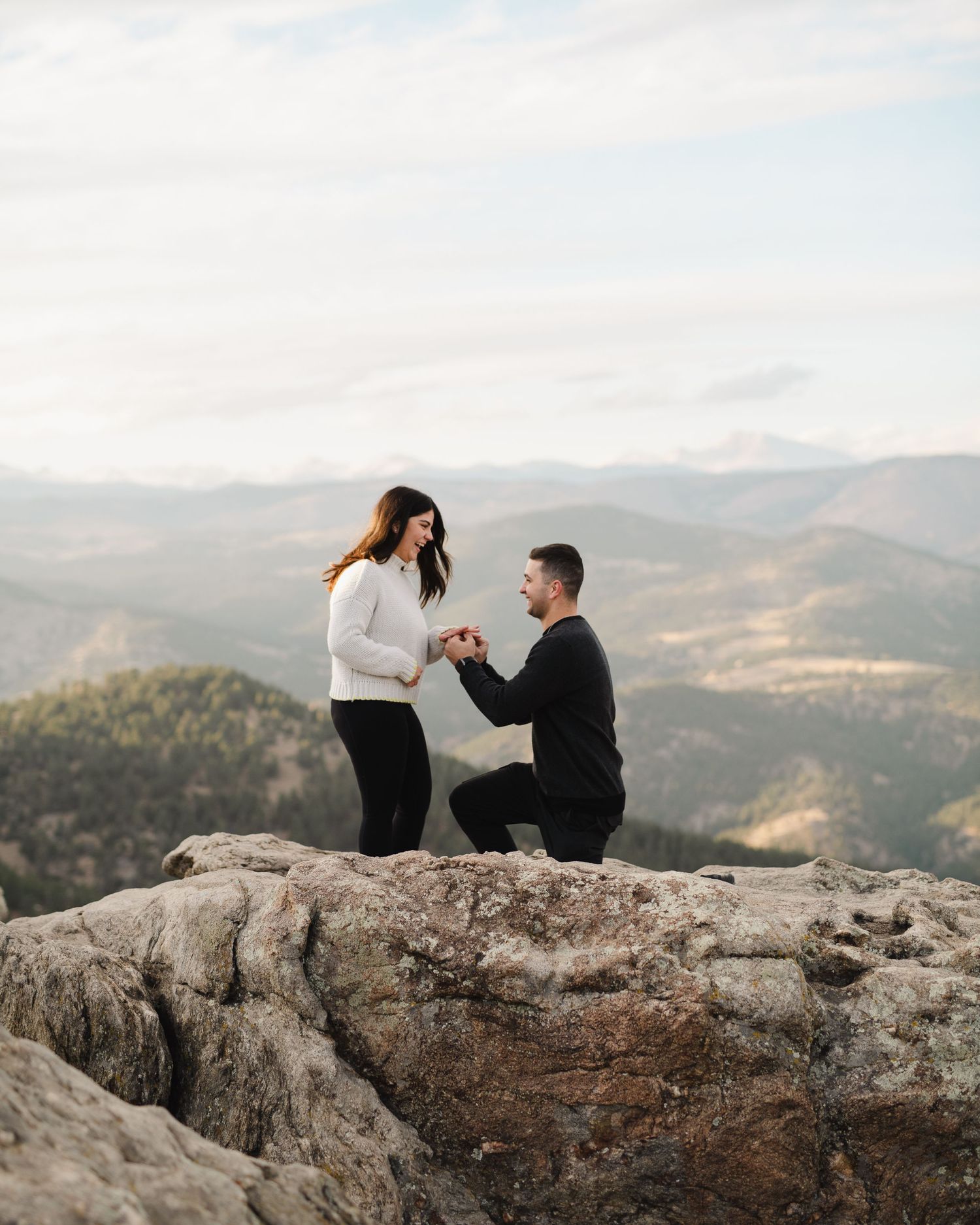 Couple shares romantic proposal moment on rocky mountain overlook with scenic valley views.