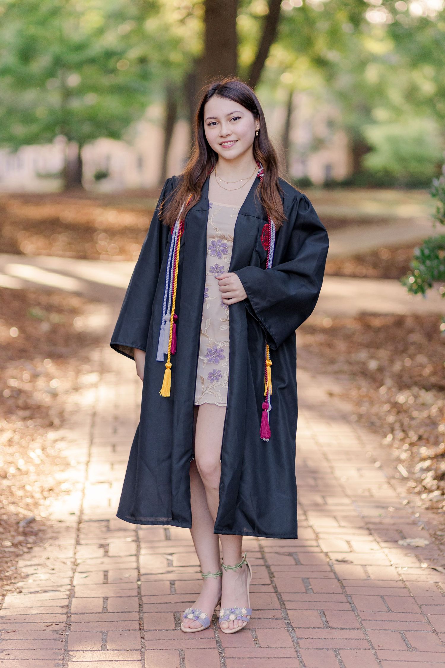 Graduate in black academic regalia stands on brick pathway surrounded by autumn trees.