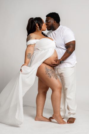 Intimate maternity photo shoot featuring a couple in white outfits against a neutral background.