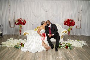 Wedding couple seated on elegant white couch with red floral arrangements and candles.