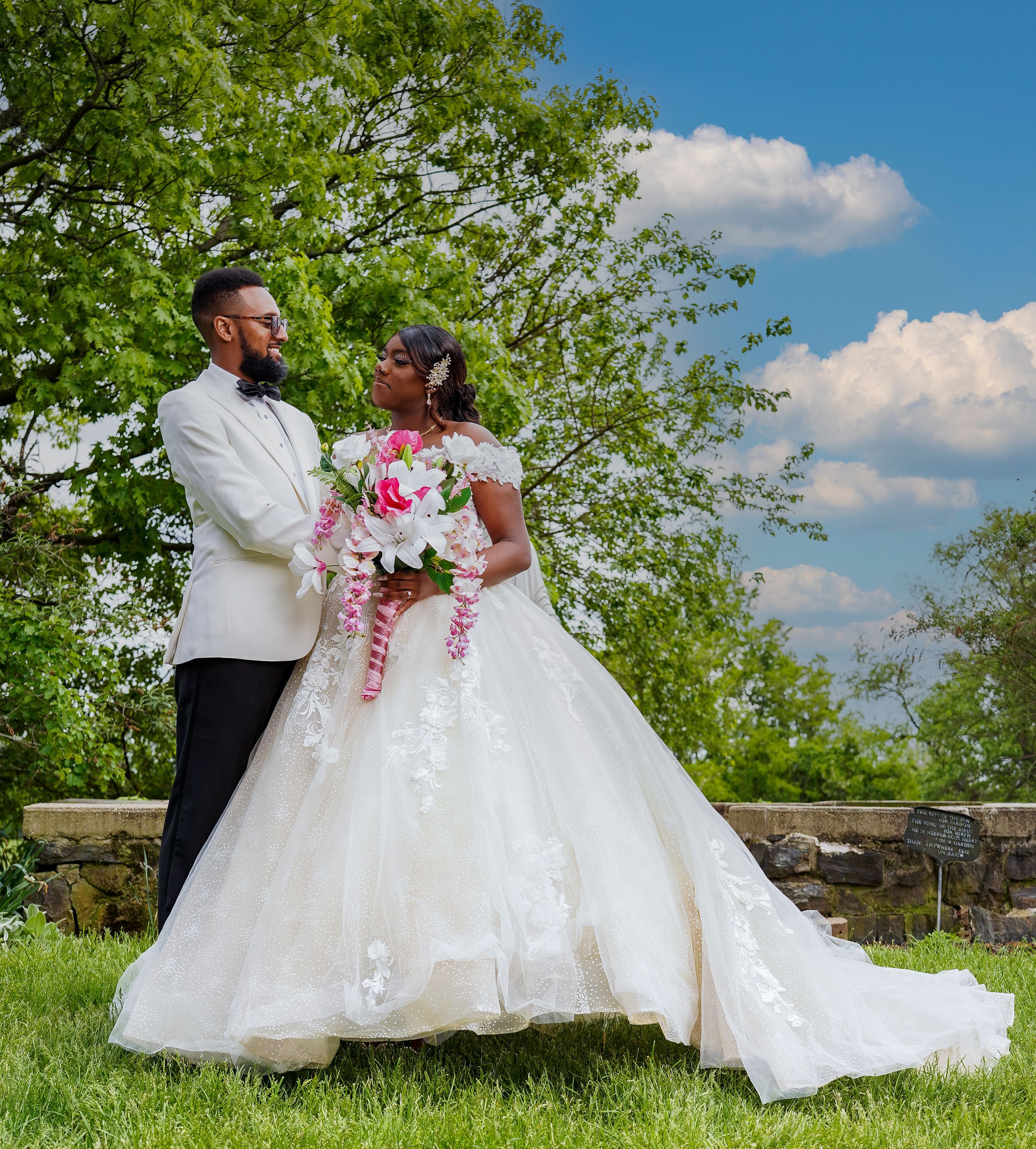 Outdoor wedding photo with couple in formal attire posing against blue sky and green trees.