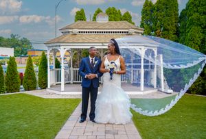 Wedding couple standing in front of white gazebo with flowing veil in garden setting.