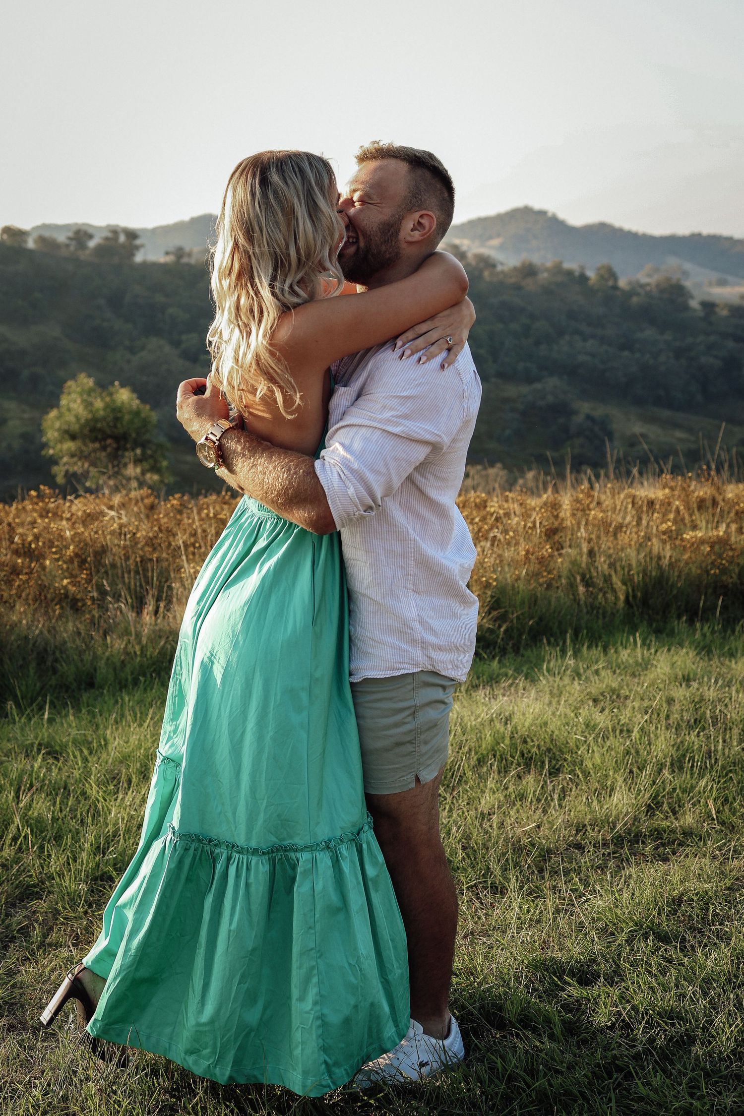 A romantic couple embraces in a grassy field at sunset with mountains in the background.