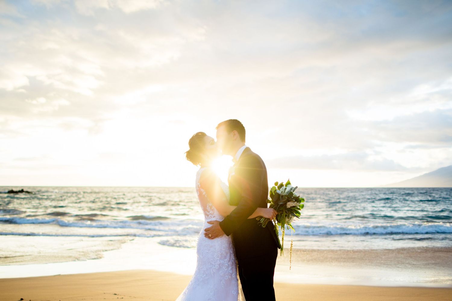 A silhouetted couple embraces on the beach during golden hour with sunlight streaming between them.