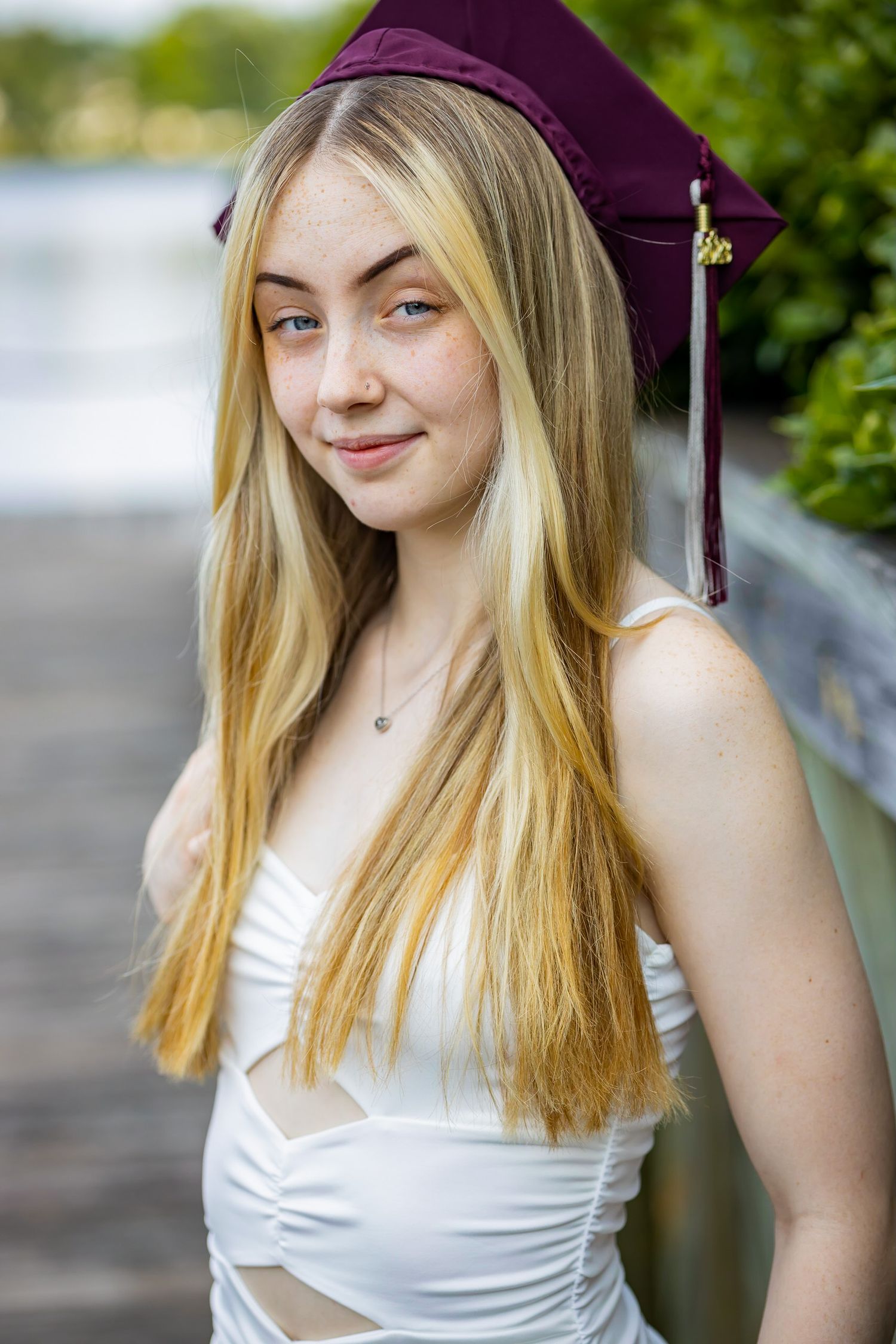 A graduate in a maroon cap and white dress poses on a wooden boardwalk for graduation photos.
