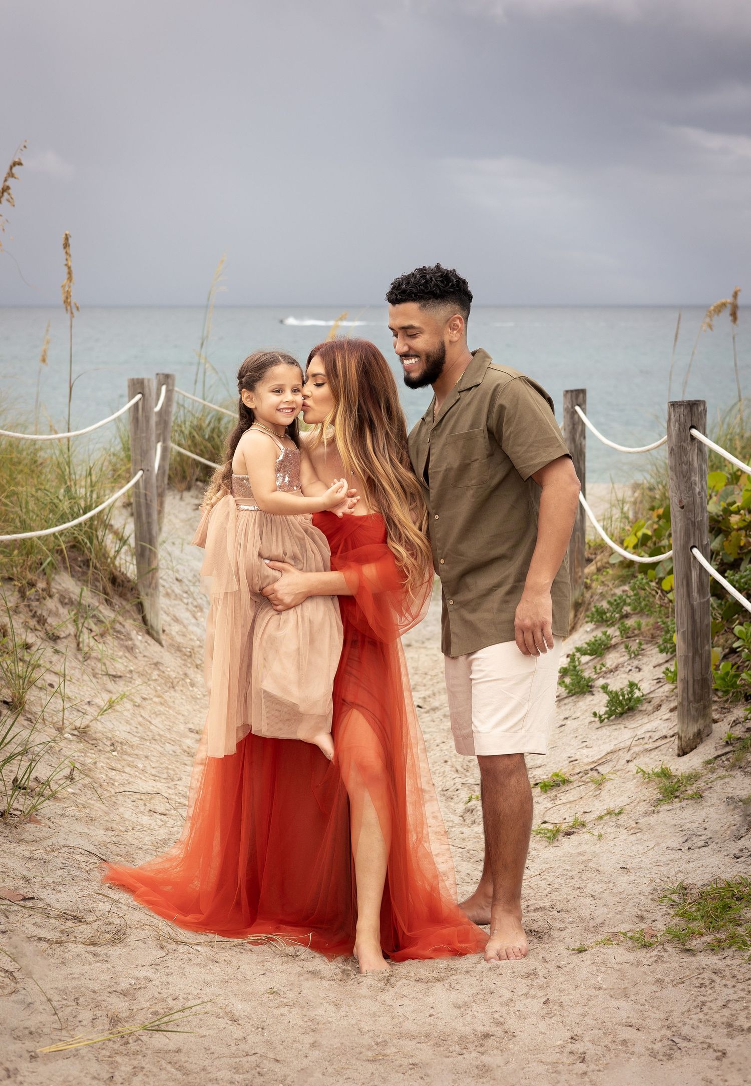 Family sharing a tender moment together on a sandy beach path with ocean and cloudy sky in background.