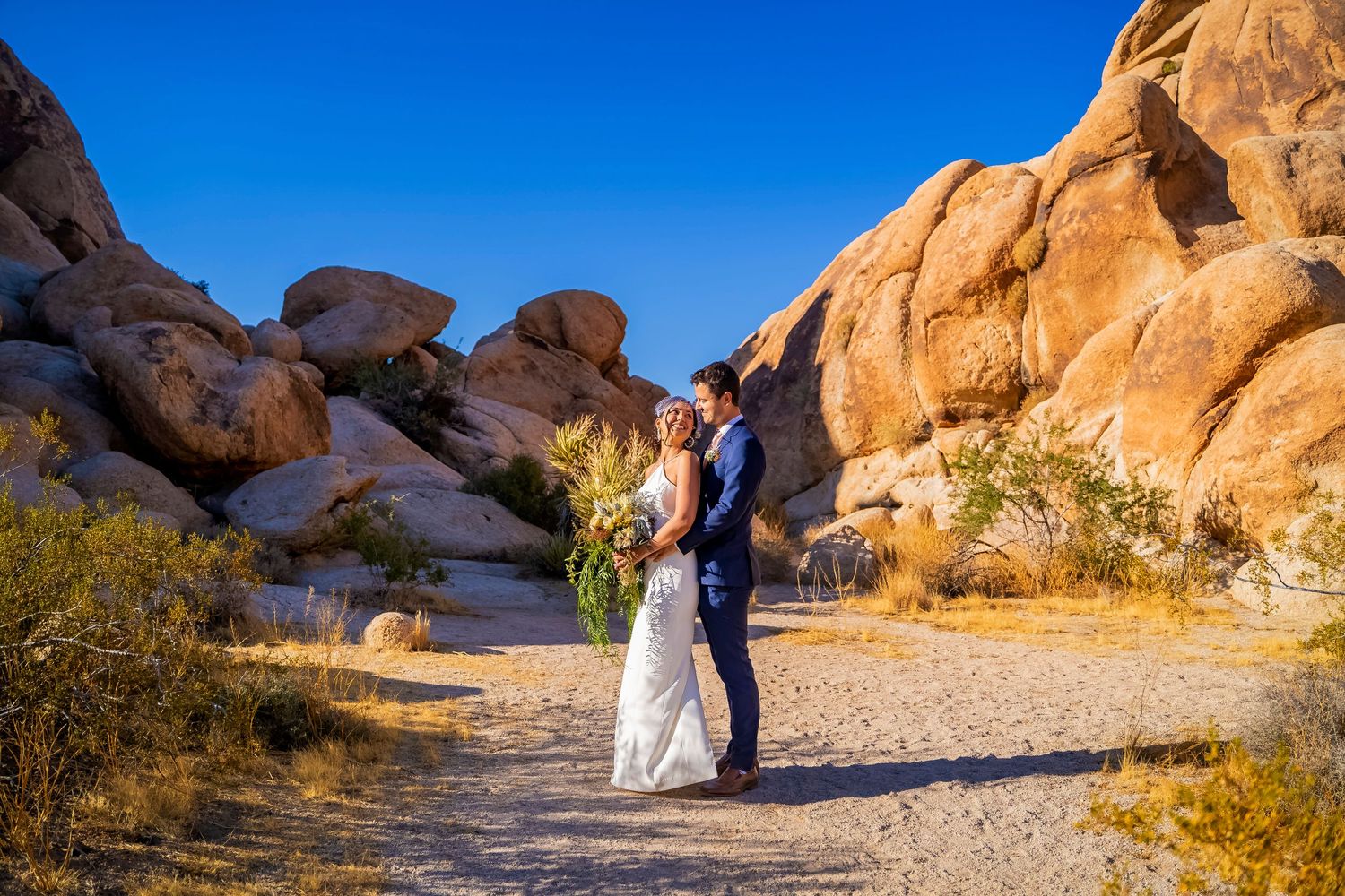 A couple in wedding attire embracing among dramatic red rock formations in a desert landscape.