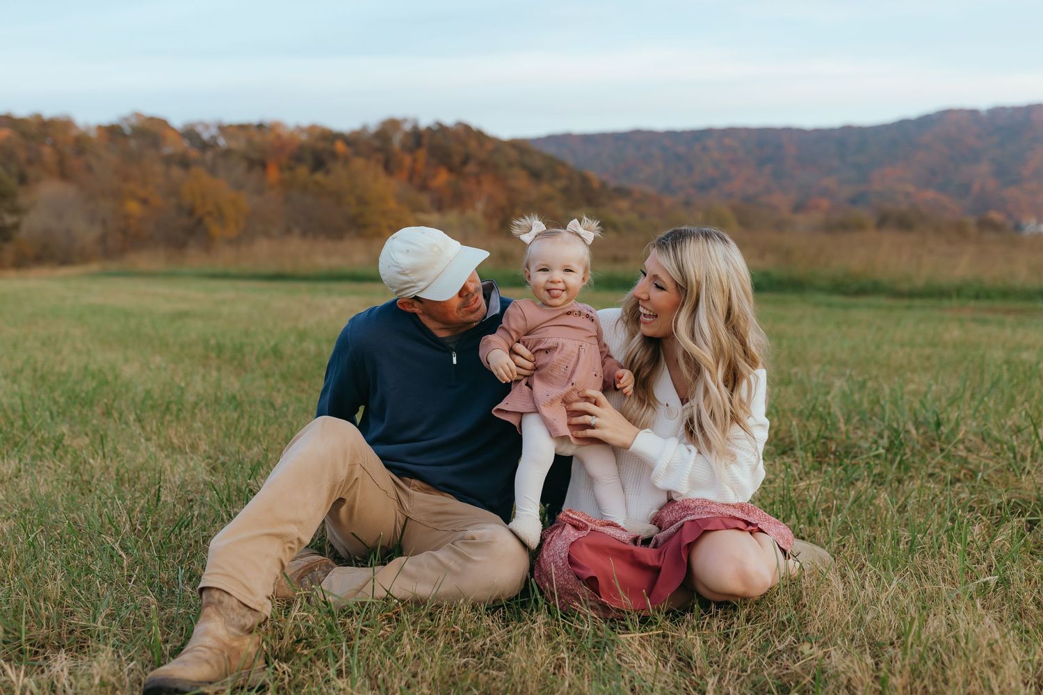 Family sits together in autumn field with mountains and fall foliage in background.