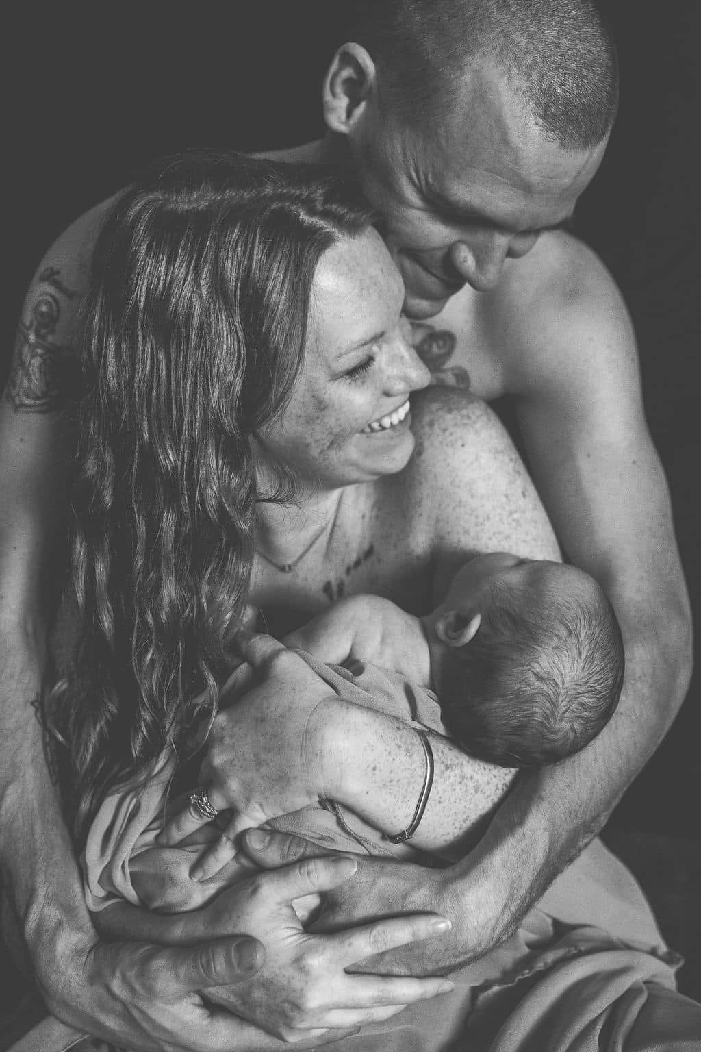 Black and white photo of smiling parents holding their newborn baby in an intimate family portrait.