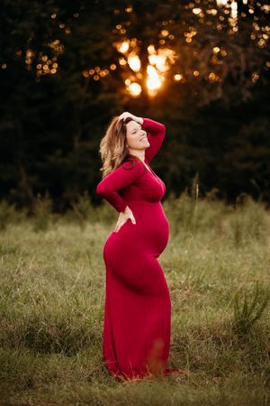 Pregnant woman in red dress posing outdoors at golden sunset, maternity photography in a lush green field.