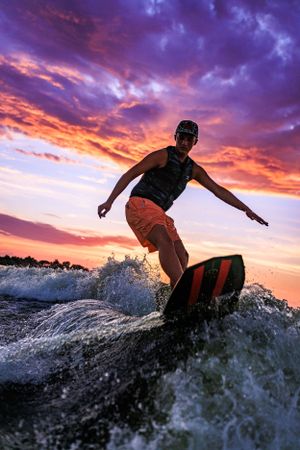Wakeboarder rides wave silhouetted against dramatic purple and orange sunset sky over lake.