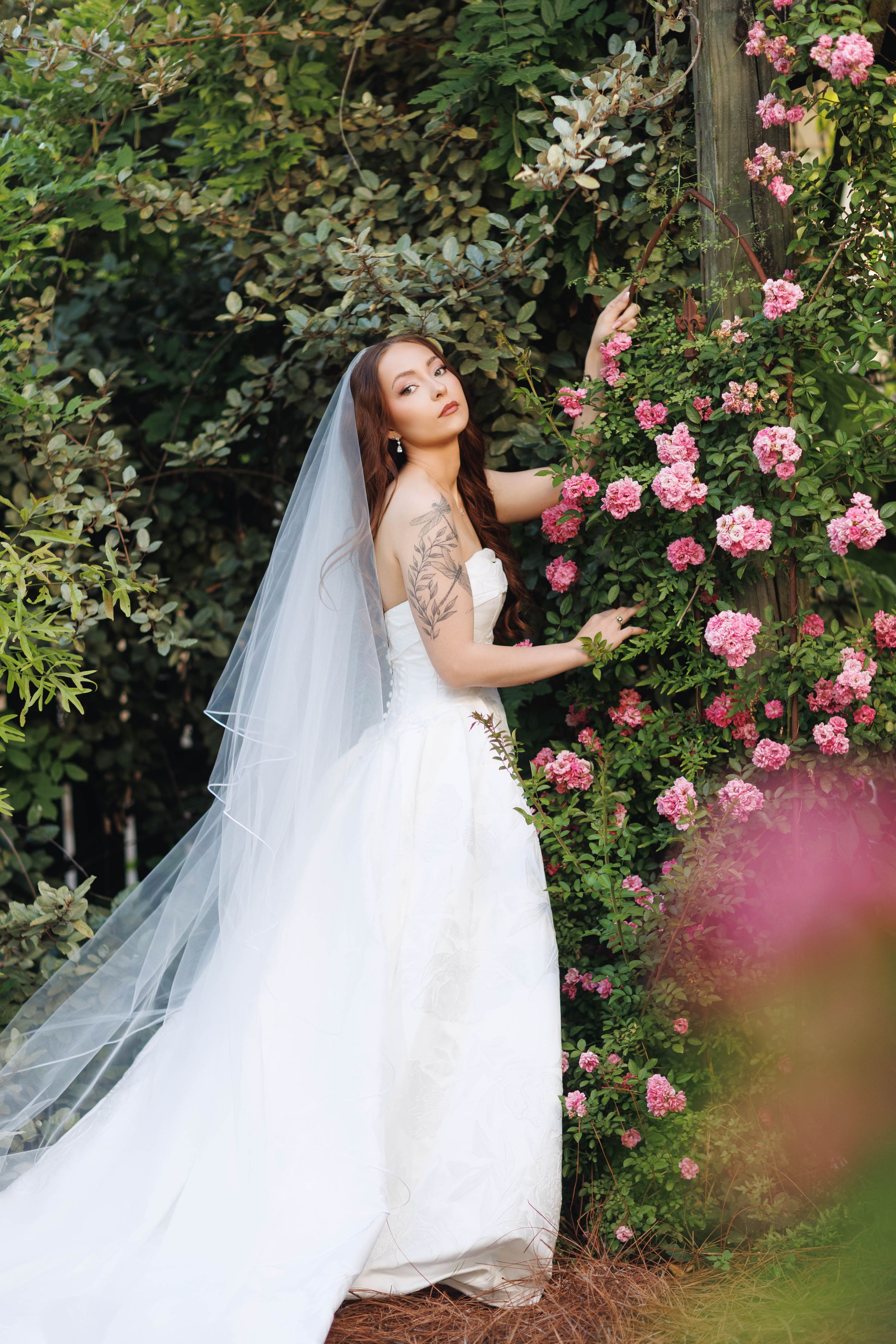 Bride in white off-shoulder gown and long veil stands beside pink roses in a lush garden.