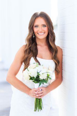 A beautiful bride in a white strapless wedding dress holds a bouquet of white roses and greenery.